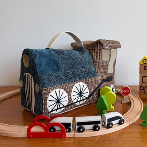 Puede incluir: Un juego de tren de juguete con una funda de transporte de tela. La locomotora es azul y marrón con detalles de ruedas blancas. La vía de tren de madera incluye vagones, árboles y una señal de stop.