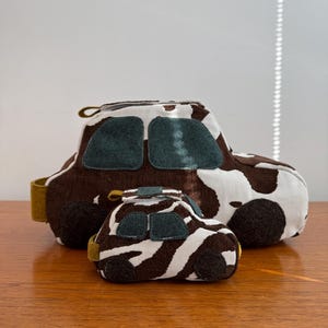 May include: Two toy cars, one large and one small, sit on a wooden surface. The larger car has a brown and white cow print pattern, with dark green window details. The smaller car has a brown and white zebra print pattern, with dark green window details.