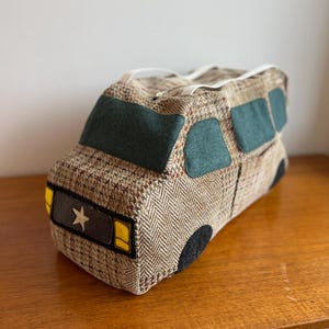 Puede incluir: Un juguete de tela con forma de furgoneta camper. La furgoneta está hecha de tela de tweed marrón con ventanas y adornos de fieltro verde. La parte delantera presenta faros amarillos y un emblema de estrella.
