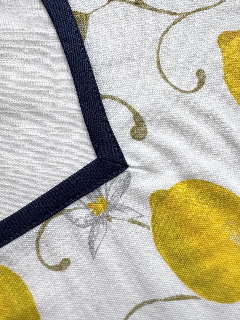 May include: White fabric with a navy blue border and a lemon and floral pattern. The design features bright yellow lemons, gray flowers, and olive green vines. The navy border frames the top left corner of the fabric.