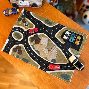 Puede incluir: Una alfombra de juego de carretera de fieltro hecha a mano con un diseño de asfalto negro, hierba verde y coches de juguete en miniatura. También hay un camión de remolque de juguete de tela y un camión de juguete azul. La alfombra presenta un diseño de ciudad y carretera.