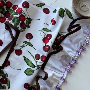 May include: White fabric with a repeating pattern of red cherries and green leaves. The fabric has a brown trim and is held together with purple clips. A metal ruler and a ceramic bowl are in the background.