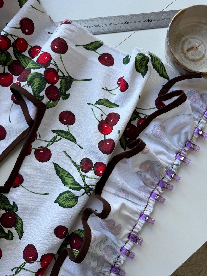 May include: White fabric with a repeating pattern of red cherries and green leaves. The fabric has a brown trim and is held together with purple clips. A metal ruler and a ceramic bowl are in the background.