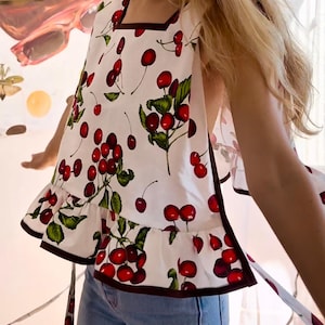 May include: White sleeveless top with a cherry print. The top features a square neckline, a ruffled hem, and dark brown trim. The design includes red cherries, green leaves, and stems on a white background.