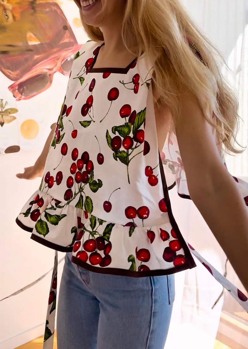 May include: White sleeveless top with a cherry print. The top features a square neckline, a ruffled hem, and dark brown trim. The design includes red cherries, green leaves, and stems on a white background.