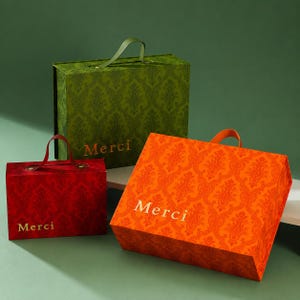 May include: Three rectangular gift boxes with handles, featuring a damask pattern in green, red, and orange. Each box displays the word Merci in gold lettering on the front. The boxes vary in size and are arranged in a cluster on a flat surface.