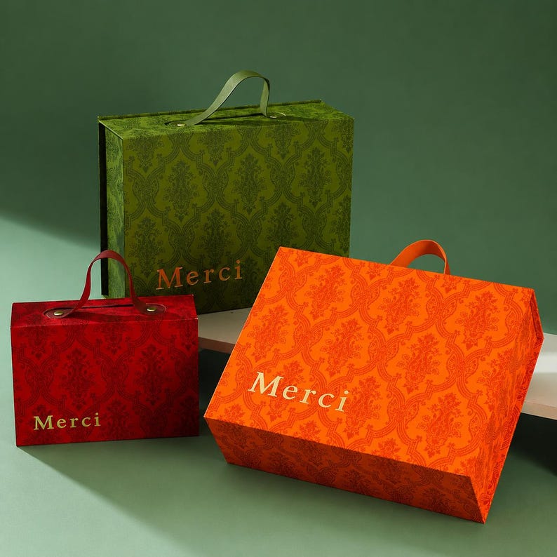 May include: Three rectangular gift boxes with handles, featuring a damask pattern in green, red, and orange. Each box displays the word Merci in gold lettering on the front. The boxes vary in size and are arranged in a cluster on a flat surface.