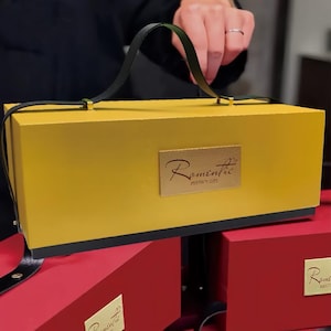 Pode incluir: Uma caixa de presente retangular amarela com uma alça e base pretas. A caixa tem uma etiqueta retangular dourada com a palavra "Romantic" e a frase "Perfect Gift". A caixa é segurada por uma pessoa.