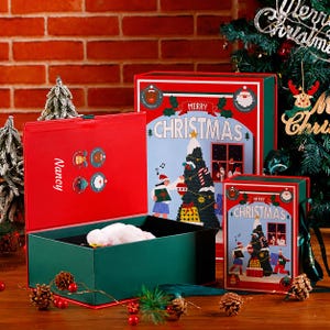 Könnte beinhalten: Eine rechteckige Geschenkbox mit rotem Deckel und dunkelgrünem Boden. Die Innenseite des Deckels zeigt den Namen Nancy und vier runde Symbole. Die Außenseite ist mit einer Frohe Weihnachten Illustration mit Baum und Figuren bedruckt.
