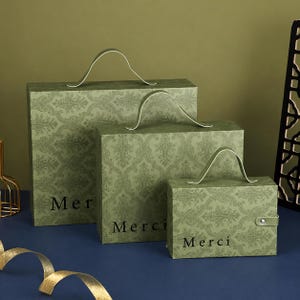 May include: Three rectangular sage green gift boxes of varying sizes, each featuring a dark green damask pattern and a top handle. The word Merci is printed in black on the front of each box. The smallest box includes a side snap closure.