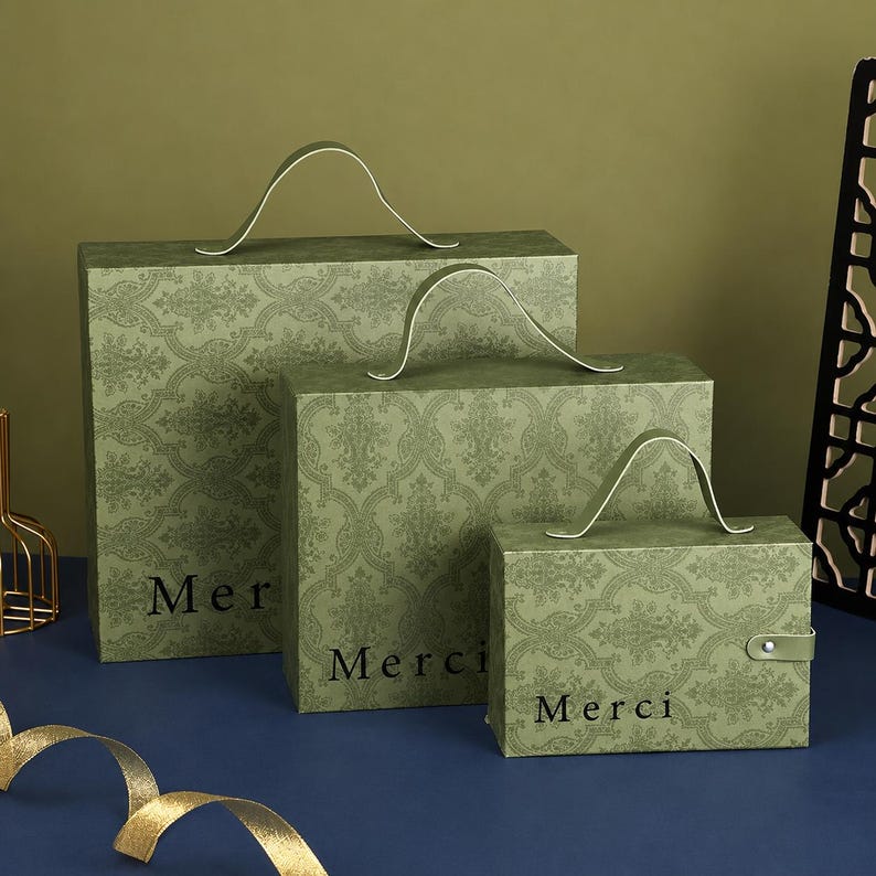 May include: Three rectangular sage green gift boxes of varying sizes, each featuring a dark green damask pattern and a top handle. The word Merci is printed in black on the front of each box. The smallest box includes a side snap closure.