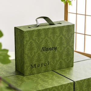 May include: A rectangular olive green gift box with a dark green damask pattern, featuring a top handle and the text "Nancy" and "Merci" printed in black on the front. The box has a structured design with a hinged lid and a smooth finish.