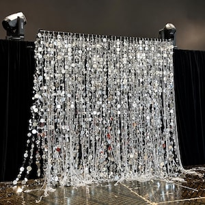 May include: A shimmering silver sequin curtain backdrop. The curtain is made of numerous strands of silver sequins, creating a dazzling effect. The backdrop is set against a black background, with stage lights on either side.