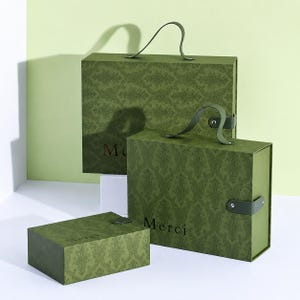 May include: Three rectangular olive green gift boxes with a dark green damask pattern. Each box features the word Merci printed in a dark font. The two larger boxes have matching green handles and side closures, while the smallest box is flat.