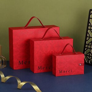 May include: Three rectangular red gift boxes of varying sizes, each featuring a top handle, a subtle damask-like pattern, and the word Merci printed in black on the front. The smallest box includes a single snap button closure on the side.