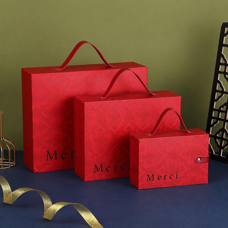 May include: Three rectangular red gift boxes of varying sizes, each featuring a top handle, a subtle damask-like pattern, and the word Merci printed in black on the front. The smallest box includes a single snap button closure on the side.