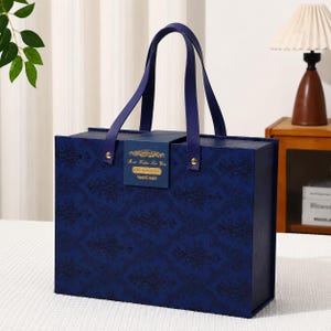 Puede incluir: Una caja de regalo rectangular de color azul oscuro con un patrón de damasco negro repetitivo. La caja cuenta con dos asas azul oscuro sujetas con remaches dorados y un cierre magnético central que muestra el texto dorado Best Wishes For You.