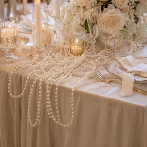 Puede incluir: Un arreglo de mesa con un mantel de tela color crema drapeado con largas tiras de perlas blancas artificiales. El conjunto incluye candelabros de vidrio transparente con velas encendidas, cristalería y un centro de mesa de rosas blancas.