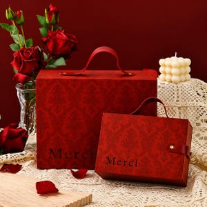 May include: Two rectangular red gift boxes with a dark red damask pattern and a top handle. Each box features the word "Merci" printed in black on the front. The boxes are constructed with a flap closure secured by a single gold-colored metal stud.