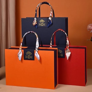 Puede incluir: Tres bolsas de regalo en naranja, rojo y azul marino, cada una con un borde oscuro contrastante. Las bolsas tienen asas de bufanda de seda y una etiqueta que dice "Forever Love". Las bolsas están dispuestas en una composición en capas.