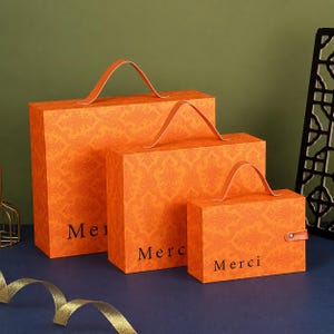 May include: Three rectangular orange gift boxes of varying sizes, each featuring a repeating damask pattern and a top handle. The word Merci is printed in black on the front of each box. The smallest box includes a side strap with a metal snap closure.