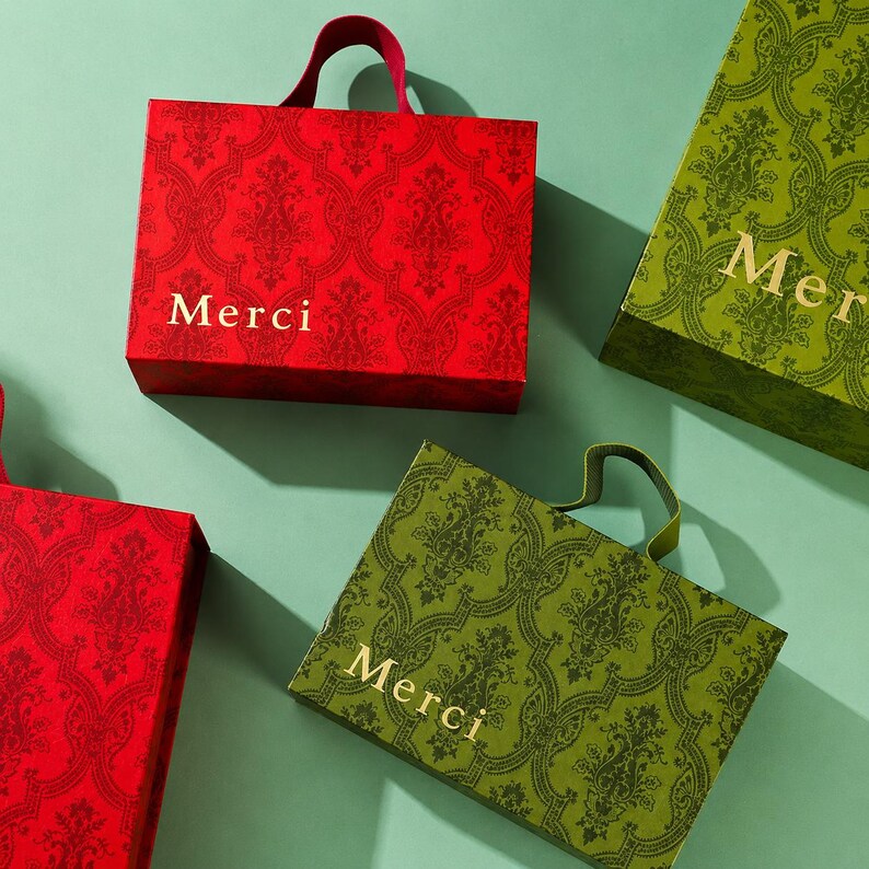 May include: Rectangular gift bags with handles, featuring a repeating damask pattern. Two bags are red with gold text reading Merci, and two bags are olive green with gold text reading Merci. The bags are arranged in a group on a flat surface.
