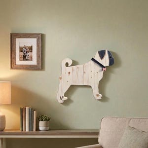 May include: A wooden pug dog wall decoration. The dog is made of light-coloured wood with a black and grey head, a black collar, and a pink bow tie. The pug is mounted on a light green wall. A framed photo hangs on the wall.