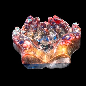 Op de afbeelding: Een glassculptuur van twee handen die bij elkaar zijn gevouwen, met een rode, blauwe en gouden gespikkelde afwerking.