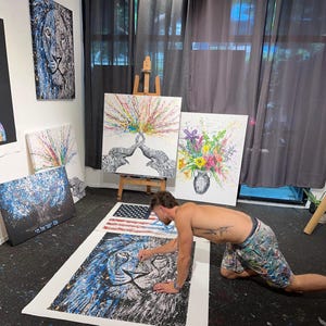 以下が含まれることがあります： スタジオにいる男性が、ライオンの顔を描いた大きなキャンバスの絵画に取り組んでいます。キャンバスは床に置かれ、男性は床にひざまずいて、手でキャンバスに絵の具を塗っています。ライオンの顔は青、白、黒の色合いで描かれています。男性は短パンを履いており、背中にタトゥーがあります。スタジオの壁には、ゾウの絵、花の絵、木の絵など、他の絵画も飾られています。