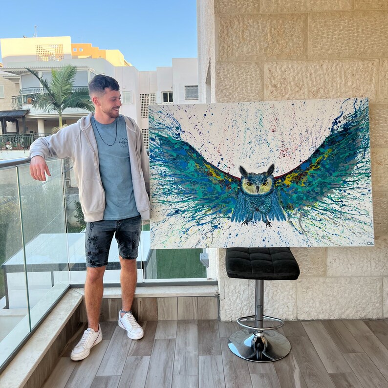 May include: A large abstract painting of an owl with blue, green, and yellow colors on a white background. The owl is in the center of the painting and has its wings spread out. The painting is hanging on a wall in a room with a person standing in front of it.