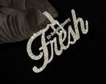 May include: A silver pendant with the word "Fresh" spelled out in cursive and covered in clear rhinestones. The pendant is held in a gloved hand.