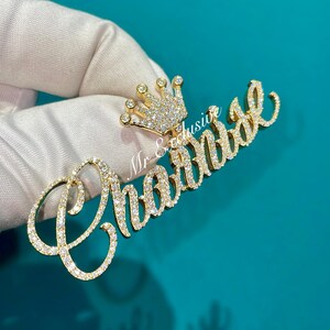 May include: A gold-colored pendant with the name "Chantel" spelled out in cursive script, encrusted with sparkling gemstones. A small crown, also adorned with gems, sits above the name. The pendant is held against a teal background.