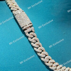 May include: Close-up of a gold-toned cuban link chain necklace. The chain is encrusted with numerous small, sparkling stones. The clasp is rectangular and also covered in stones. The background is a solid teal color.