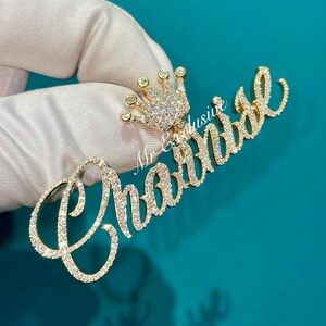 May include: A gold-colored pendant with the name "Chantal" written in a cursive font, encrusted with small, clear gemstones. A small crown, also covered in gemstones, sits above the name. The pendant is held against a teal background.