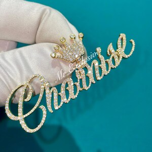 May include: A gold-colored pendant with the name "Chantal" in cursive script, encrusted with small, sparkling stones. A small crown, also adorned with stones, sits above the name. The pendant is held against a teal background.