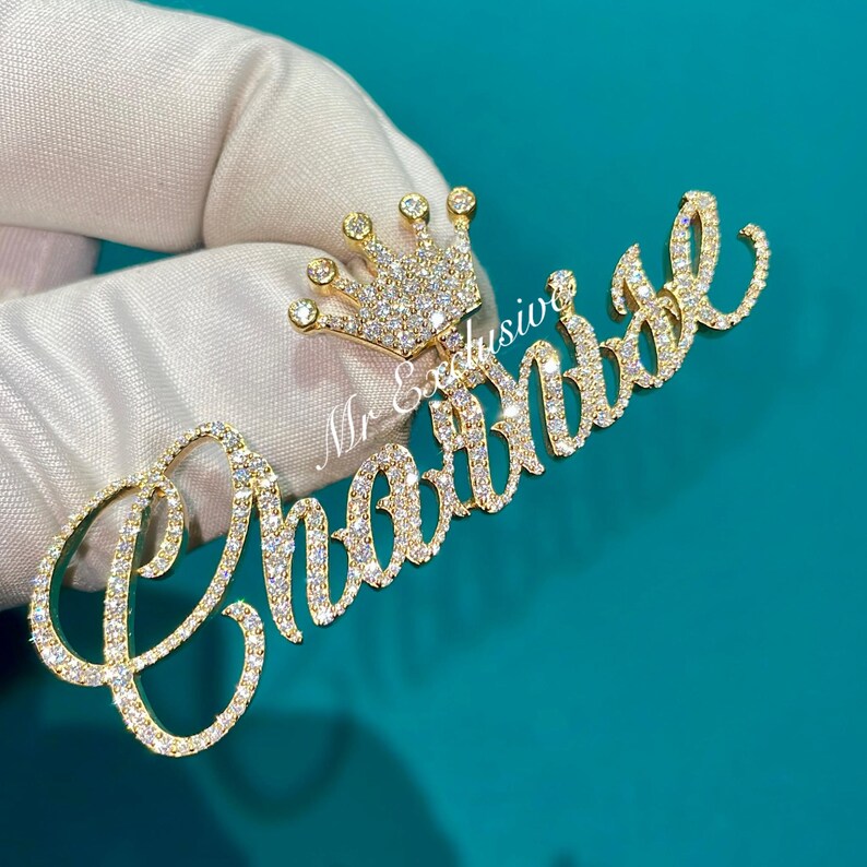 May include: A gold-colored pendant with the name "Chantal" in cursive script, encrusted with small, sparkling stones. A small crown, also adorned with stones, sits above the name. The pendant is held against a teal background.