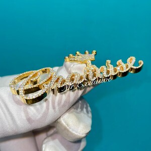 May include: A gold-colored pendant with the name "Chanise" in a cursive font, encrusted with small, clear gemstones. A small crown is above the name. The words "Mr. Exclusive" are below the name. The pendant is held by a gloved hand.
