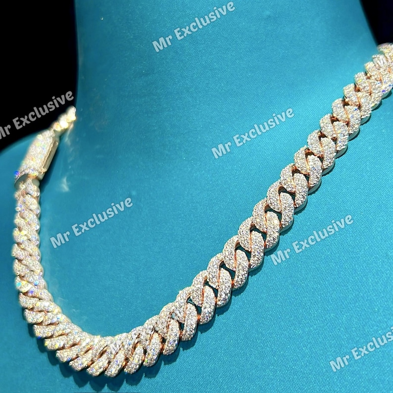 May include: A gold-toned, iced-out Cuban link chain necklace. The necklace features a series of interlocking, rounded links, each encrusted with small, sparkling stones. The clasp is also detailed with similar stones. The background is a solid teal color.