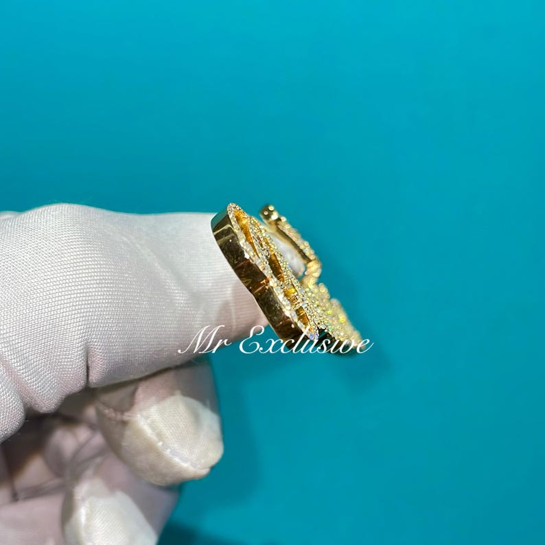 May include: A gold-colored pendant with a detailed design, encrusted with small, sparkling stones. The pendant is held against a white fabric with the text "Mr Exclusive" visible.