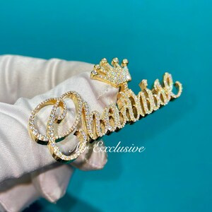 May include: A gold-colored pendant with the name "Chantal" spelled out in cursive script, encrusted with small, sparkling gemstones. A small crown is positioned above the "t". The pendant is held against a white surface.