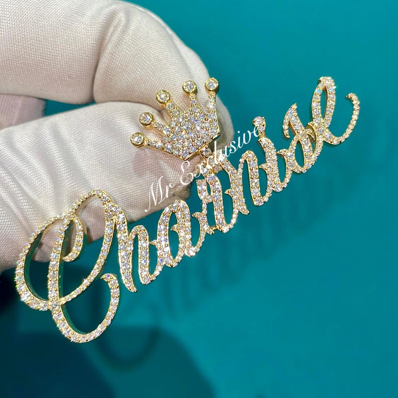 May include: A gold-toned pendant with the name "Charnise" in a cursive font, encrusted with small, clear gemstones. A small crown, also adorned with gemstones, sits above the name. The pendant is held against a teal background.