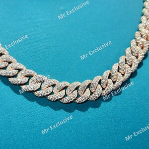 May include: Rose gold cuban link chain necklace encrusted with sparkling white gemstones. The necklace is displayed against a teal background. The chain links are closely interlocked, creating a bold and luxurious look.