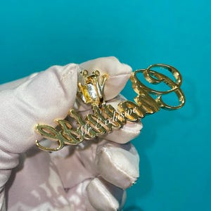 May include: A gold-colored pendant with the text "Mr Exclusive" in a cursive font. The pendant has a crown design and a large letter "B" at the end. The pendant is held by a gloved hand.