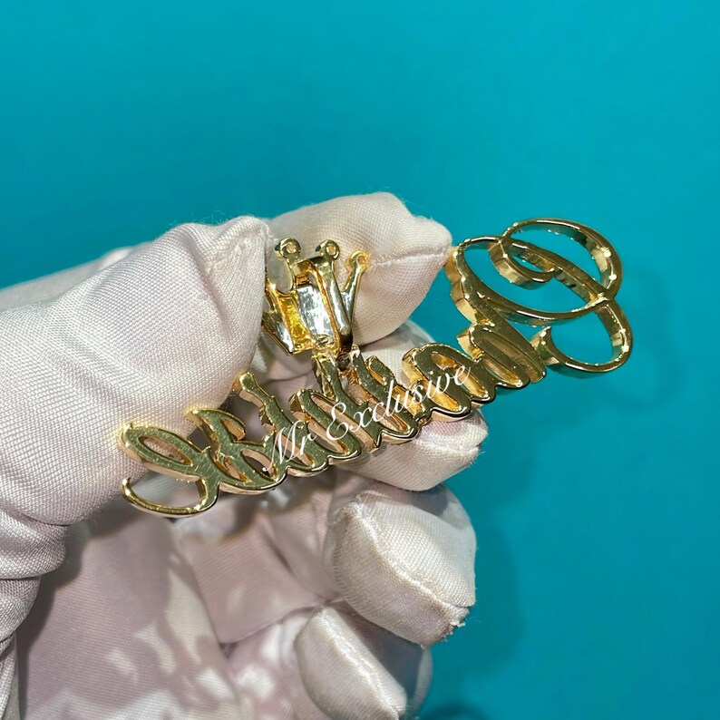 May include: A gold-colored pendant with the text "Mr Exclusive" in a cursive font. The pendant has a crown design and a large letter "B" at the end. The pendant is held by a gloved hand.