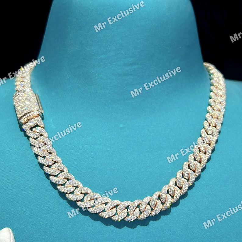 May include: A gold-toned cuban link chain necklace encrusted with sparkling, clear gemstones. The necklace has a secure clasp and is displayed on a teal background. The text "Mr Exclusive" is visible.