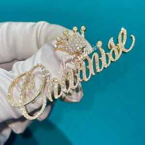 May include: A gold-colored pendant with the name "Chanel" spelled out in cursive, encrusted with clear gemstones. A small crown, also adorned with gemstones, sits above the name. The text "Mr Exclusive" is visible.