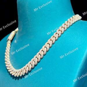 May include: A gold-toned cuban link chain necklace, encrusted with numerous small, sparkling, clear stones. The necklace is displayed on a blue surface, showcasing its intricate design and luxurious appearance.