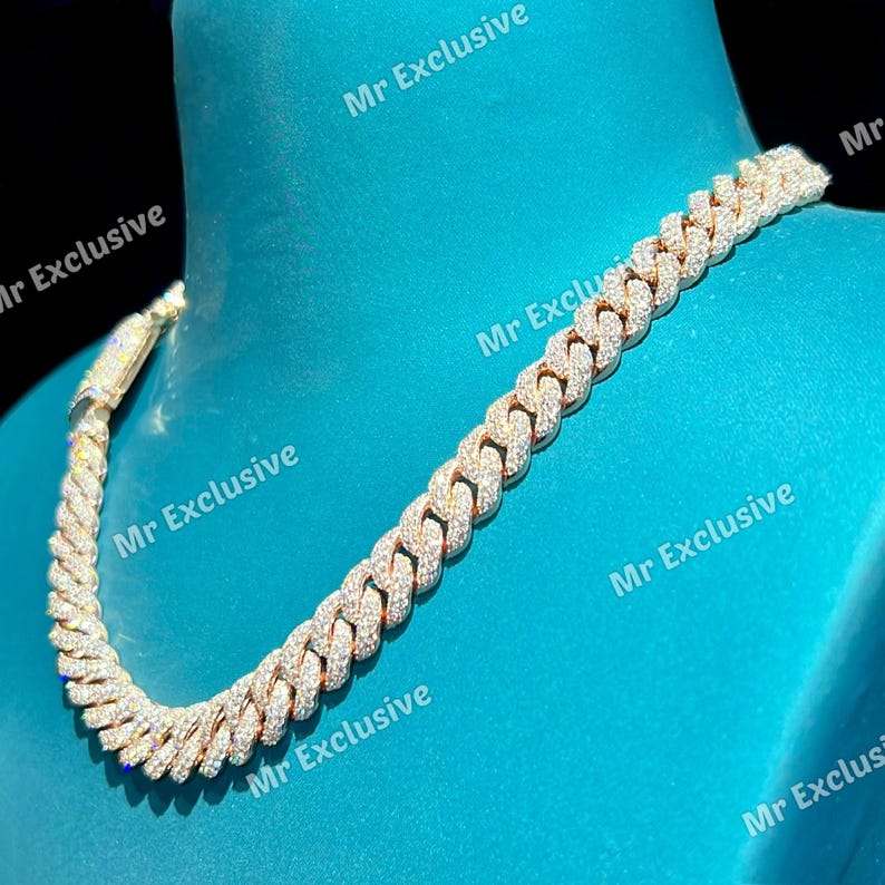 May include: A gold-toned cuban link chain necklace, encrusted with numerous small, sparkling, clear stones. The necklace is displayed on a blue surface, showcasing its intricate design and luxurious appearance.