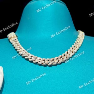 May include: A gold-toned cuban link chain necklace encrusted with numerous small, clear gemstones. The necklace is displayed on a blue surface. The text "Mr Exclusive" is visible in the image.