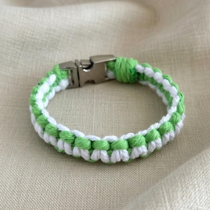 Peut inclure: Un bracelet tressé avec des bandes alternées de cordon blanc et vert vif. Le bracelet est doté d'un fermoir en métal argenté. L'accessoire est présenté sur un tissu de couleur neutre. Circonférence d'environ 18 cm.